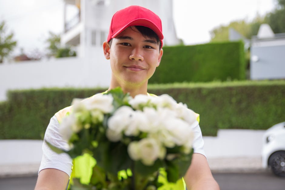 A delivery person wearing a red cap and a high-visibility yellow vest is holding a bouquet of fresh white flowers, including roses and freesias, wrapped in clear plastic with a white ribbon around the