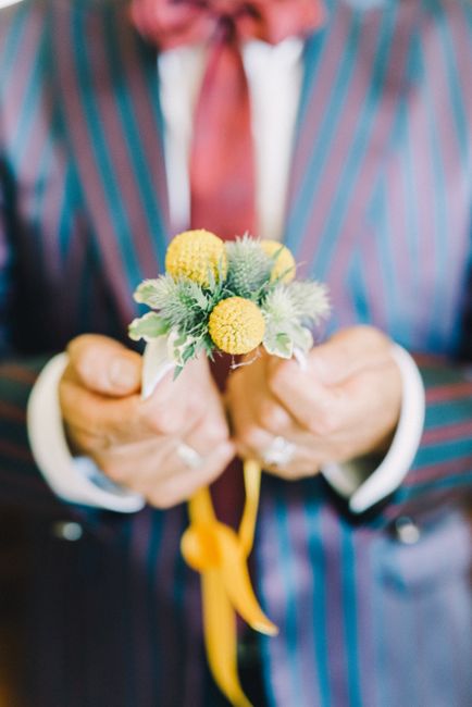 A man dressed in a pinstripe suit with a white shirt and red tie is holding a small floral bouquet, which features yellow button flowers, soft green foliage, and white dusty miller leaves. The bouquet