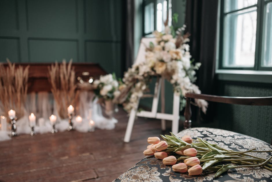A bouquet of pale peach tulips with green stems and leaves is casually placed on an ornate fabric armchair with a floral pattern, inside a softly lit room with dark green walls and large windows. In t