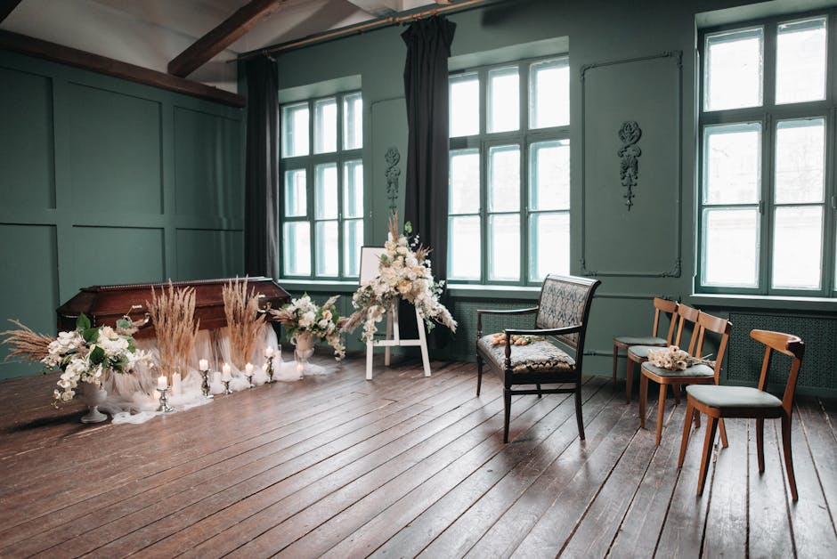 A floral arrangement featuring white roses, peonies, and delphiniums, accented with greenery, arranged in a cascading style on a white easel in a room with large windows. The flowers are set against a