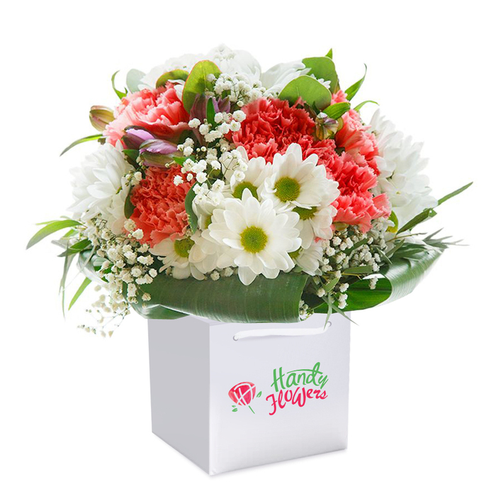 Resting atop a pure white surface, this impeccable bouquet rises proudly from a clean, square gift bag-its beauty a fitting tribute to Upper Norwood's vibrant artistry. Soft coral and blush carnations, known for their abundant, frilly petals, form a lush core, their hues softly blending with vibrant bursts of red. White daisies, accented by striking lime-green centers, add uplifting brightness that calls to mind the first blooms of spring along Jasper Road. Delicate sprays of baby's breath thread a gentle weave through the arrangement, each petite cluster adding a weightless layer of charm. Tucked subtly within are unopened purple-tinged buds, evoking the promise of new beginnings so familiar in Upper Norwood's leafy residential avenues. The skirt of the bouquet fans out with deep, shiny green leaves, curving elegantly to provide a natural wrap that enhances the vibrant blooms. Arranged in a perfect half-sphere, the bouquet is both full and balanced, its artistry evident in every carefully placed stem. Held by crisp white ribbon handles, the sturdy bag speaks of easy giving-ideal for birthdays, anniversaries, or spontaneous tokens of affection. Each detail, from colour harmony to structure, is accentuated by a soft, clean backdrop-a visual celebration of freshness and happiness, ready for delivery by the trusted hands of local Upper Norwood florists.