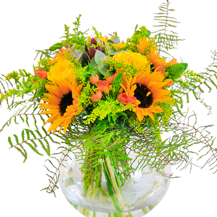 First impressions of this bouquet evoke pure sunlight, much like the golden glows often glimpsed over Upper Norwood's rooftops at dawn. Resplendent sunflowers with honeyed petals and striking, dark centers share space with sumptuous yellow garden roses, each petal spun into creamy layers. Hints of bright red hypericum berries flicker amidst the green foliage, offering gleams of contrast against the subtle matte hues of the main blooms, like autumn berries found in Grangewood Park. Fern fronds and wild, airy filler gather at the base, cascading gently and giving the bouquet a sense of movement and life. Teal-toned eucalyptus leaves slip between blossoms, infusing the composition with a fresh, earthy aroma and a cool, modern edge. Each verdant stem is visible through the vase's rounded clarity-its fishbowl shape highlighting the bouquet's balanced structure and the artistry of Upper Norwood's talented florists. Whether chosen for a birthday or an anniversary, or simply to brighten a table on a quiet afternoon, this arrangement captures a season's celebration and brings it to life within familiar local surrounds.